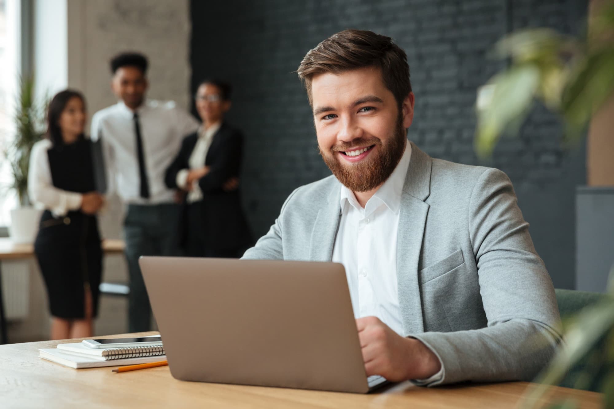 A business owner smiling at his laptop while his team talks in the background
